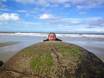 Portrait of woman hiding behind moeraki boulders at beach against sky