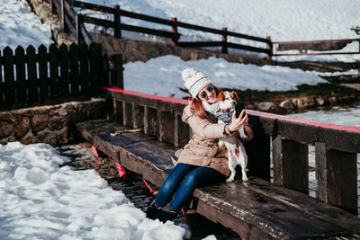 Woman taking selfie with dog while sitting on bench amidst snow covered field