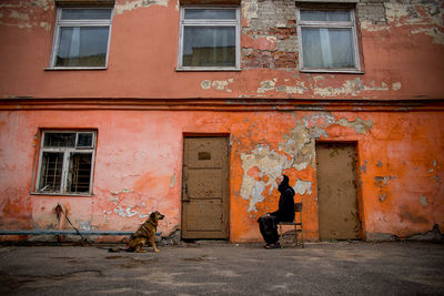 Side view of woman sitting on chair in front of dog against old building
