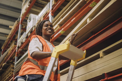 Smiling carpenter standing on ladder by rack while working in warehouse