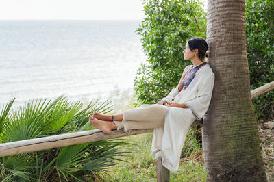 Woman looking at sea while sitting on railing near tree