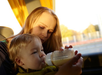 Mother feeding milk to son by window while traveling in bus