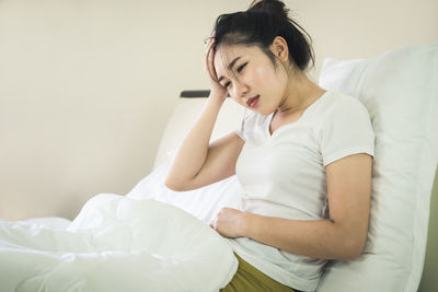 Woman with headache and stomach ache sitting on bed at home