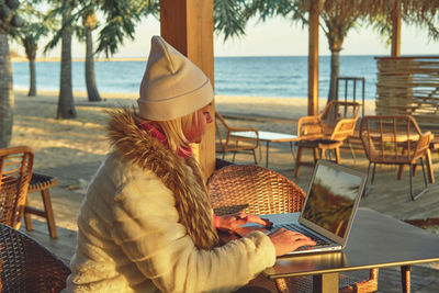 A woman in a sweater and a fur coat is sitting on the terrace of a cafe with a laptop.