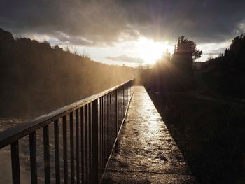 Footpath by railing against sky during sunset