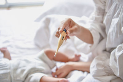 Midsection of mother holding thermometer while sitting with toddler son on bed