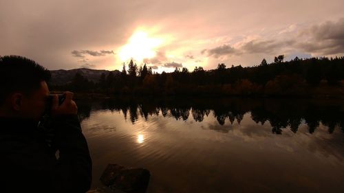 Reflection of man photographing in lake against sky during sunset