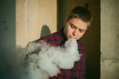 Young man smoking while standing against wall