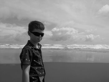 Boy wearing sunglasses standing on beach against sky black and white