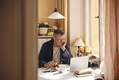 Smiling man with wired in-ear headphones watching laptop while sitting at dining table near window
