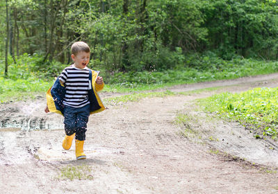 A happy little boy in a yellow jacket and rubber boots runs through the puddles and laughs. 