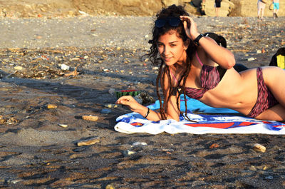 Portrait of seductive woman adjusting sunglasses while lying at beach on sunny day