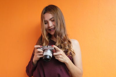 Smiling young woman photographing while standing against orange wall