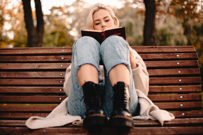 Teenage girl reading book while sitting on bench in park during autumn