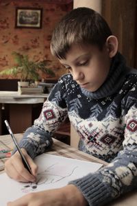 A boy in a christmas sweater sits at the table and draws and writes