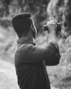 Side view of young man holding binoculars while standing outdoors