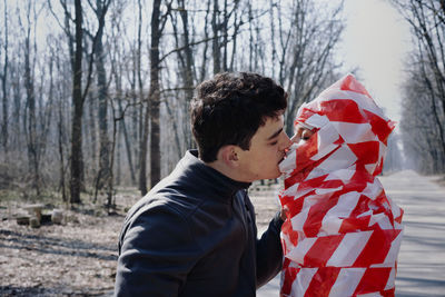 Side view of man kissing woman wrapped in cordon tape standing in forest