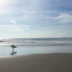 Side view of man with surfboard standing at beach against sky