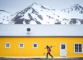 A woman walking with skis in front of a yellow building with mountains