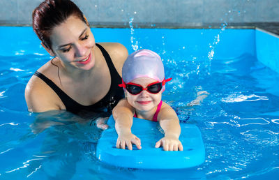 Woman teaching little girl how to swim in indoor pool with pool floating board