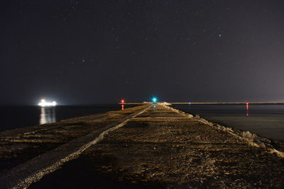 Illuminated road by sea against sky at night