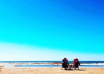 Friends sitting on deck chairs at beach against clear blue sky