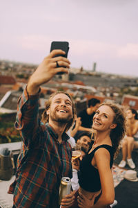 Happy man taking selfie with cheerful friends during rooftop party on terrace