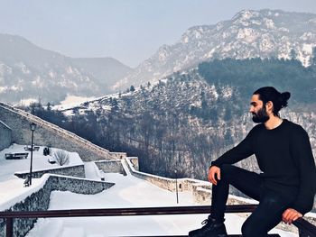 Young man sitting on railing against snowcapped mountains
