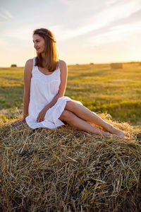 Young woman in a white dress is sitting on the haystack during a summer sunset