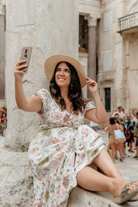 Young woman sitting on the side of a pillar taking a selfie with her mobile phone