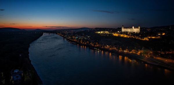 Illuminated buildings by river against sky at sunset