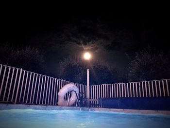 Illuminated street light by swimming pool against sky at night