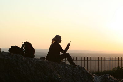 Silhouette woman smoking cigarette while sitting on rock at mountain against sky during sunset