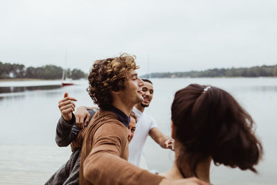 Smiling friends with arms around standing by sea against sky during vacation