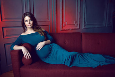 Pregnant woman in blue dress lying on red sofa in studio