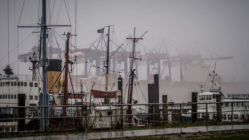 Boats by cranes against sky during foggy weather