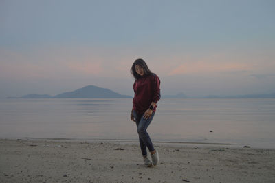 Portrait of young woman standing at beach against sky during sunset