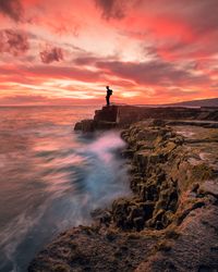 Silhouette man standing on rock formation by sea against dramatic sky during sunset