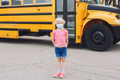 Happy caucasian girl student wearing face mask near yellow bus. new normal during coronavirus.