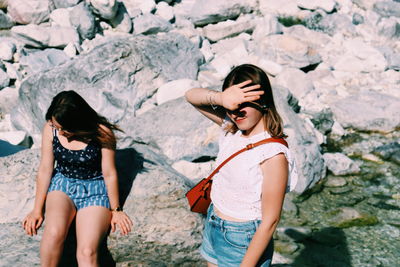 Woman shielding eyes with sister sitting on rock at beach