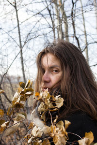 A young woman hides behind autumn leaves, wearing a black jacket with long brown hair. 