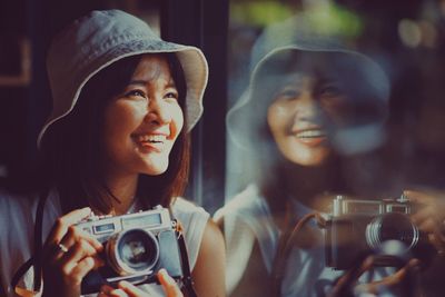 Smiling woman holding camera while leaning on glass window