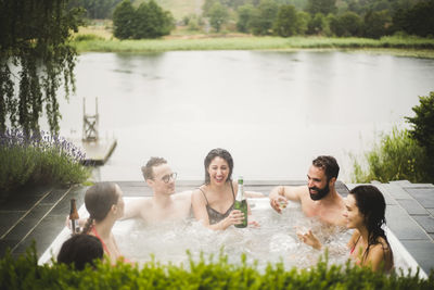 Cheerful male and female friends enjoying drinks in hot tub against lake during weekend getaway