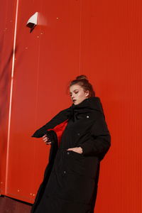 Fashionable teenage girl wearing black warm clothing standing against red wall during sunny day