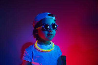 Cyberpunk boy child in white t shirt and sunglasses neon sticks on neck listening music on wall