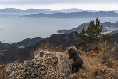 Side view of mature woman photographing sea while crouching on mountain against sky