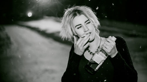 Close-up of young woman smoking cigarette while holding alcohol bottle at night