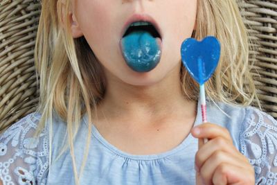 Midsection of girl sticking out tongue while holding blue heart shape lollipop