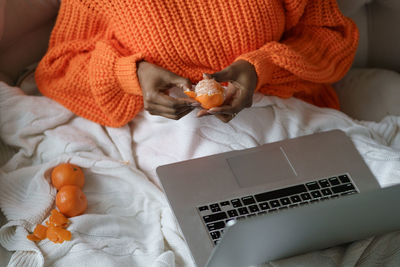 Woman hands peeling ripe tangerine, lying in bed under white plaid and working on laptop at home. 