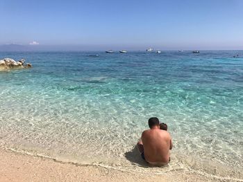 Rear view of shirtless father sitting with son on sea shore at cala mariolu